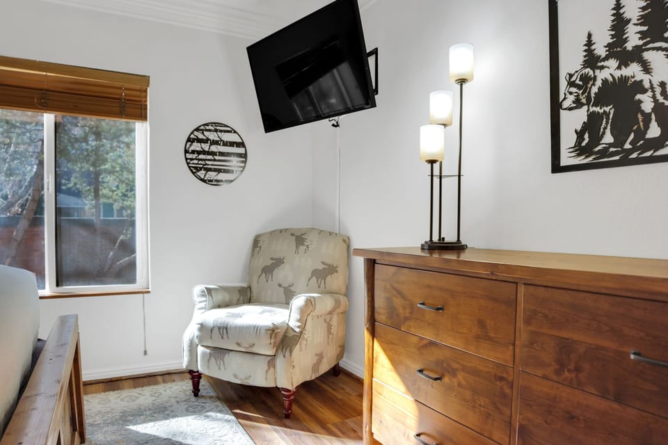 The main suite has a wood dresser, artistic table lamp, reading chair, and a flat screen wall mounted TV.