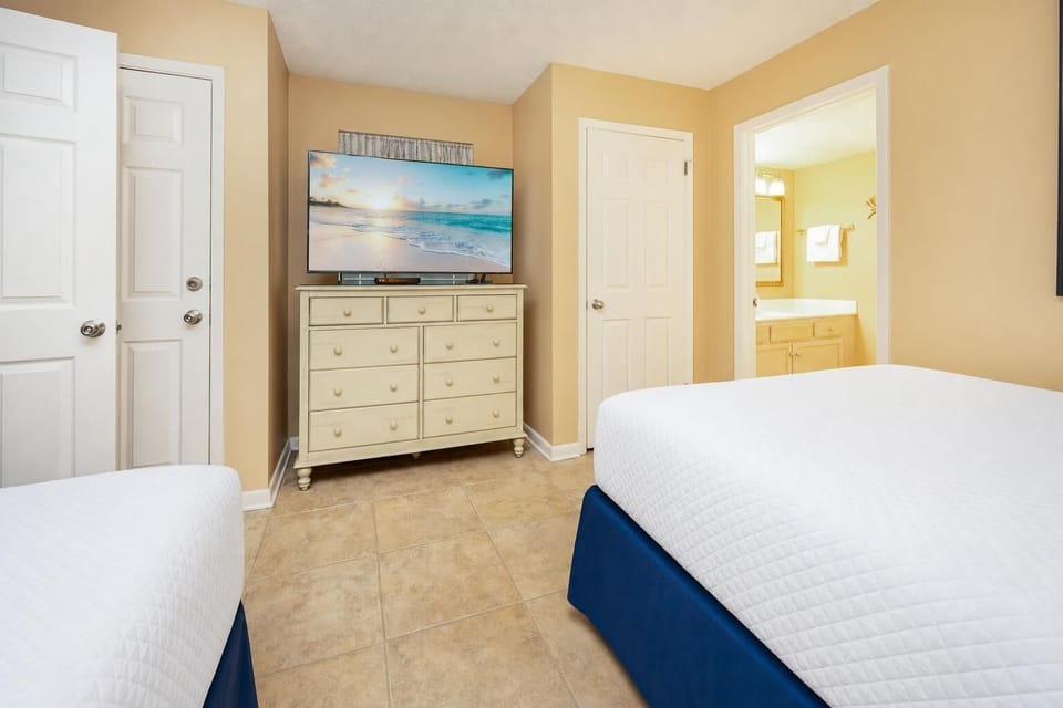 Cozy guest bedroom featuring a large TV, two beds, dresser, and a view into the bathroom with a bright, inviting atmosphere