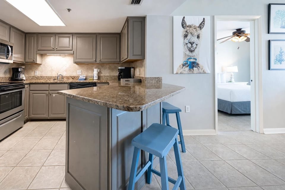 Modern kitchen features granite countertops and blue bar stools, with a whimsical llama painting adding a playful touch