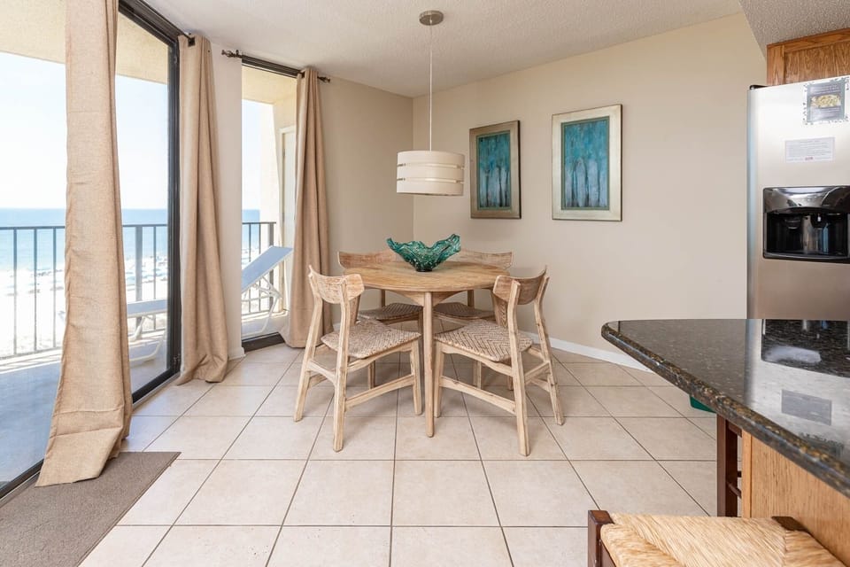 Bright dining area with a wooden table, four chairs, and a view of the beach through large sliding glass doors