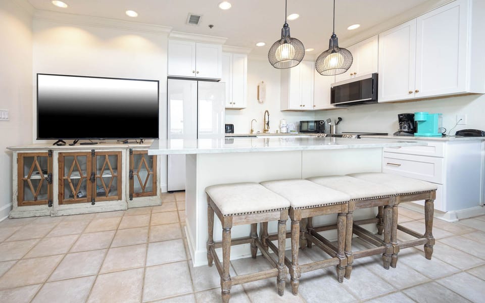Bright and airy kitchen area features a marble-topped island bar with seating, stainless appliances, and a large TV