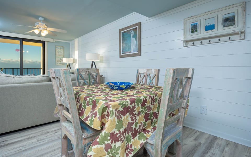 The dining area features a colorful leaf-patterned tablecloth, a blue decorative bowl, and a stunning view of the beach