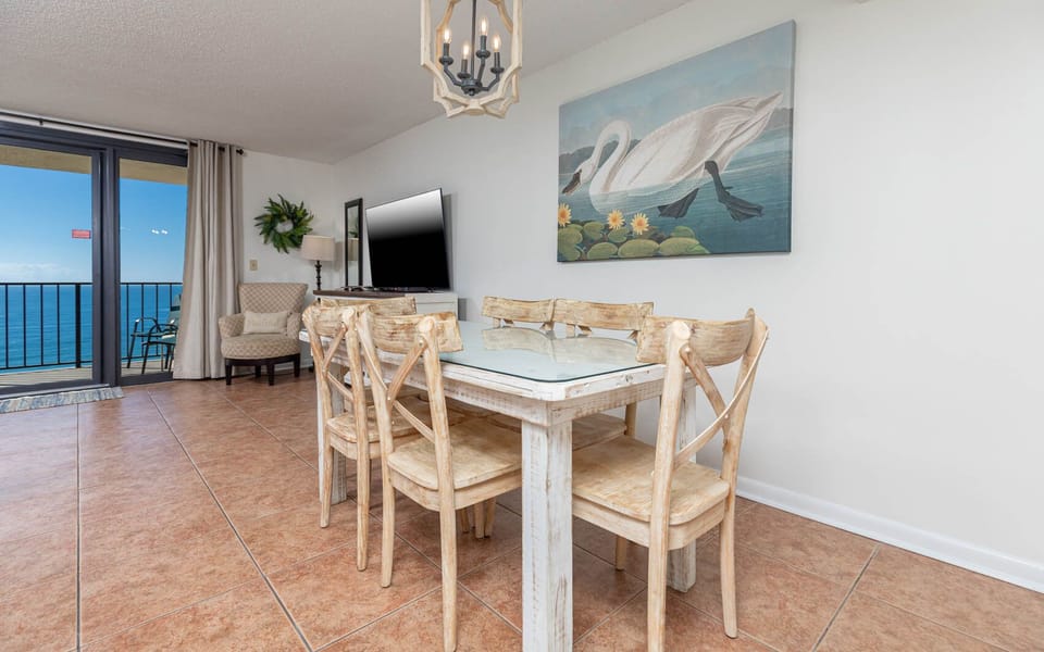 Bright dining area featuring a glass-top table, rustic chairs, and a scenic view of the tranquil waters beyond the balcony
