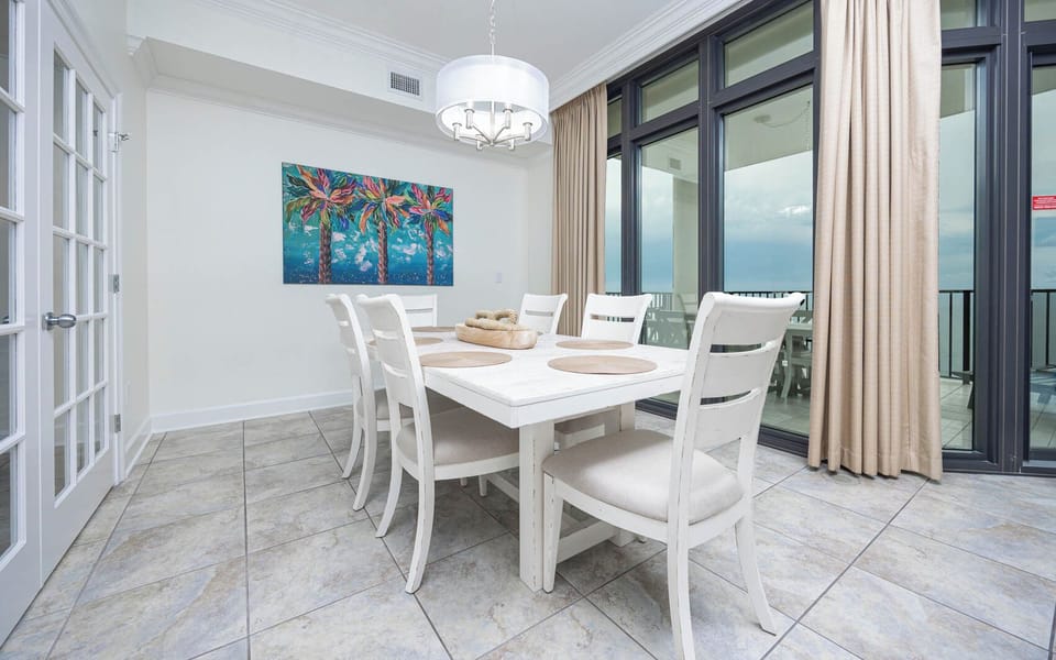 This dining area features a white table with six chairs, colorful palm art, and large windows offering a beachfront view