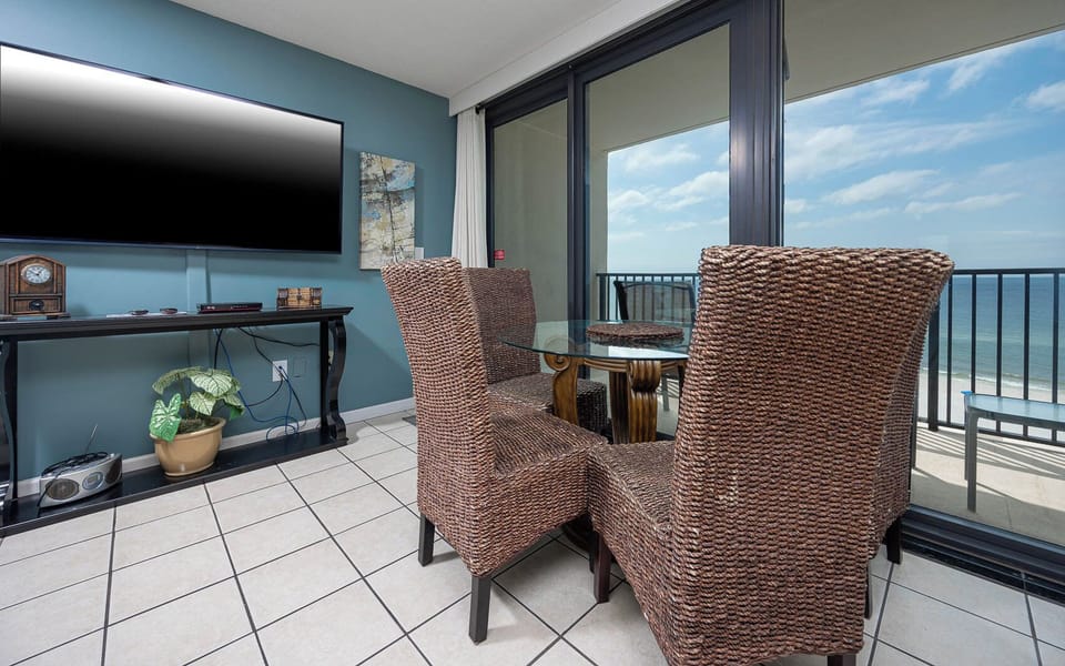 This dining area features a glass table, woven chairs, a large TV, and a stunning beachfront view through expansive windows