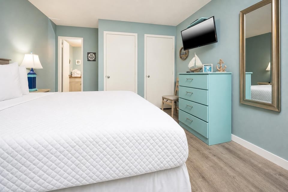 Bright and inviting guest bedroom featuring a cozy queen bed, stylish aqua dresser, and a wall-mounted TV for entertainment