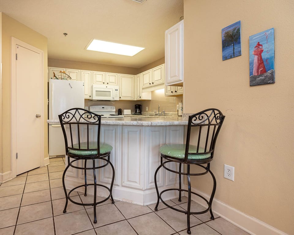 Bright kitchen area features a breakfast bar with green cushioned stools, white cabinetry, and decorative wall art