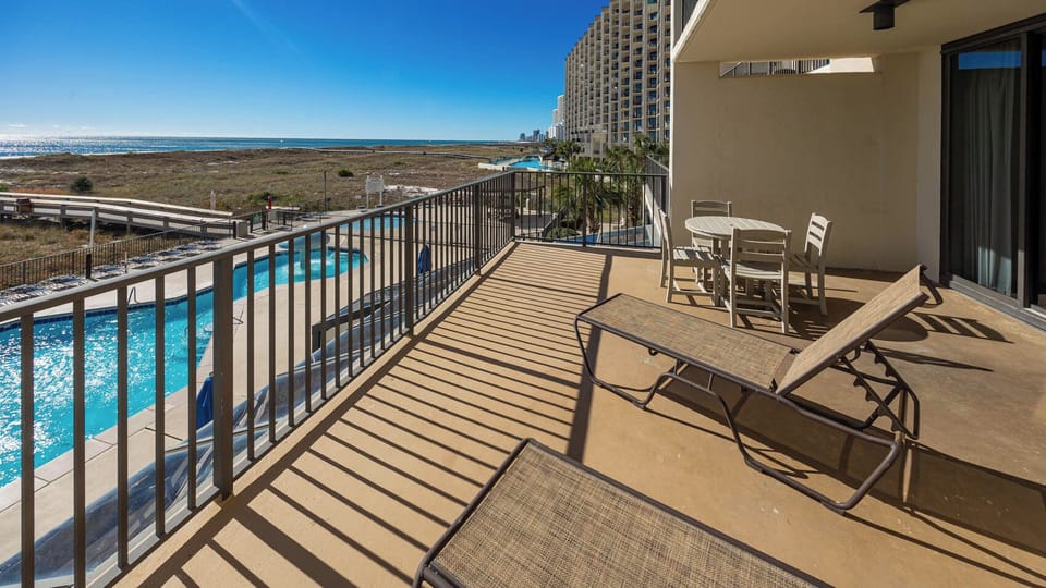 Extended and spacious balcony features loungers, patio table, and offers views of the sparkling pool area and beach beyond