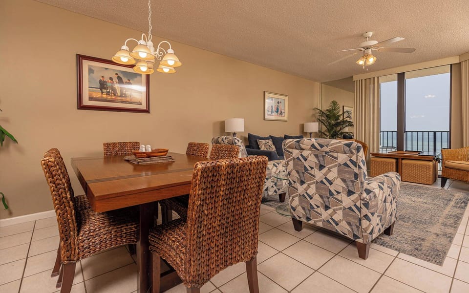 Dining table with woven chairs complements the cozy living area, featuring patterned armchairs and a view of the beach beyond