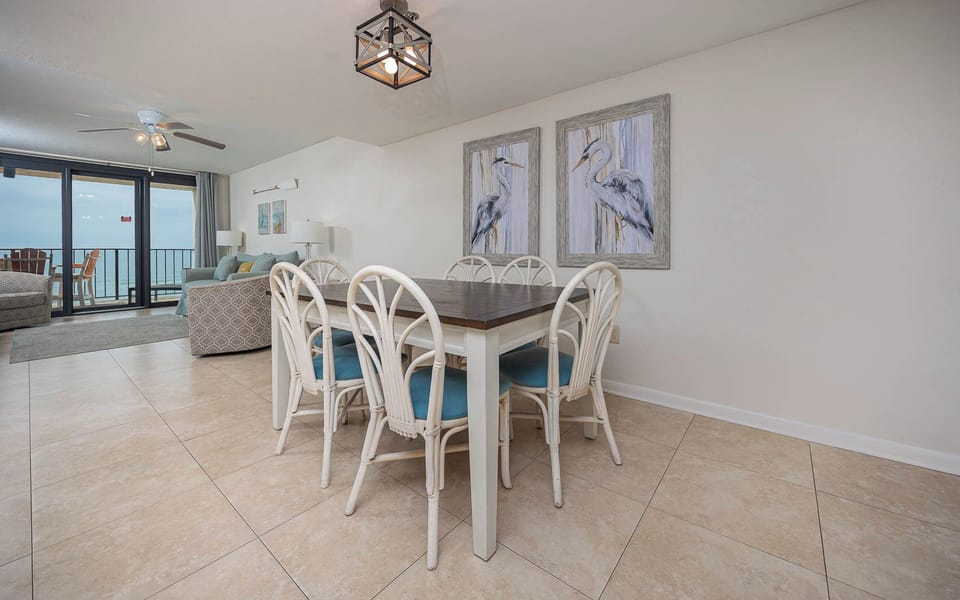 This inviting dining area features a large table with stylish chairs, complemented by coastal artwork and a view of the beach