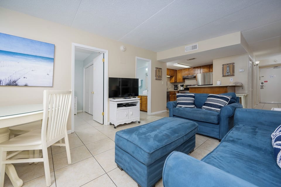 The living area features comfortable blue sofas, a dining table, and a view of the beach through the artwork