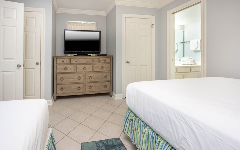 Bright guest bedroom featuring two beds, a wooden dresser with a TV, and an en-suite bathroom visible through the doorway