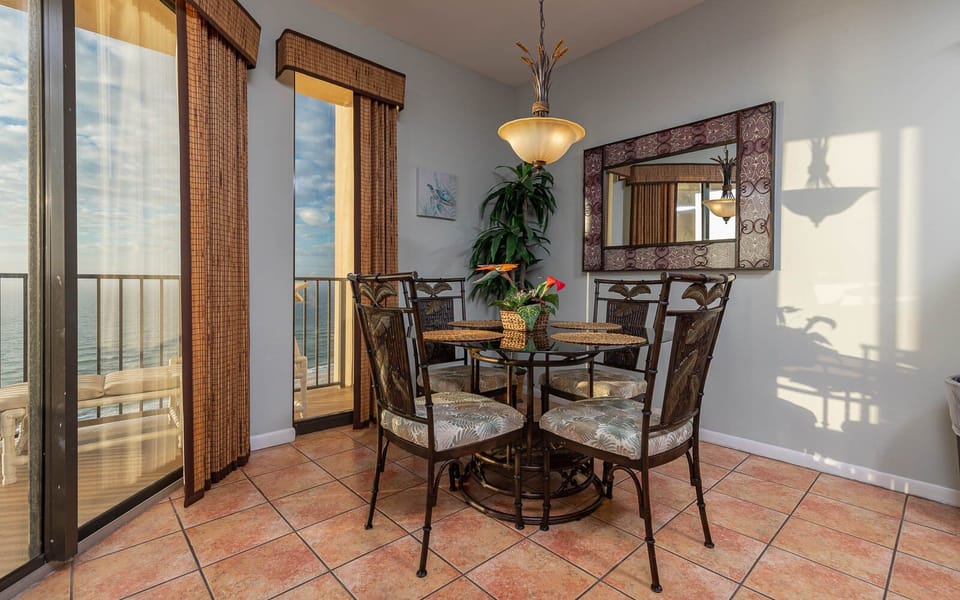 A round dining table with tropical chairs sits near large windows, offering a view of the beach and bright natural light