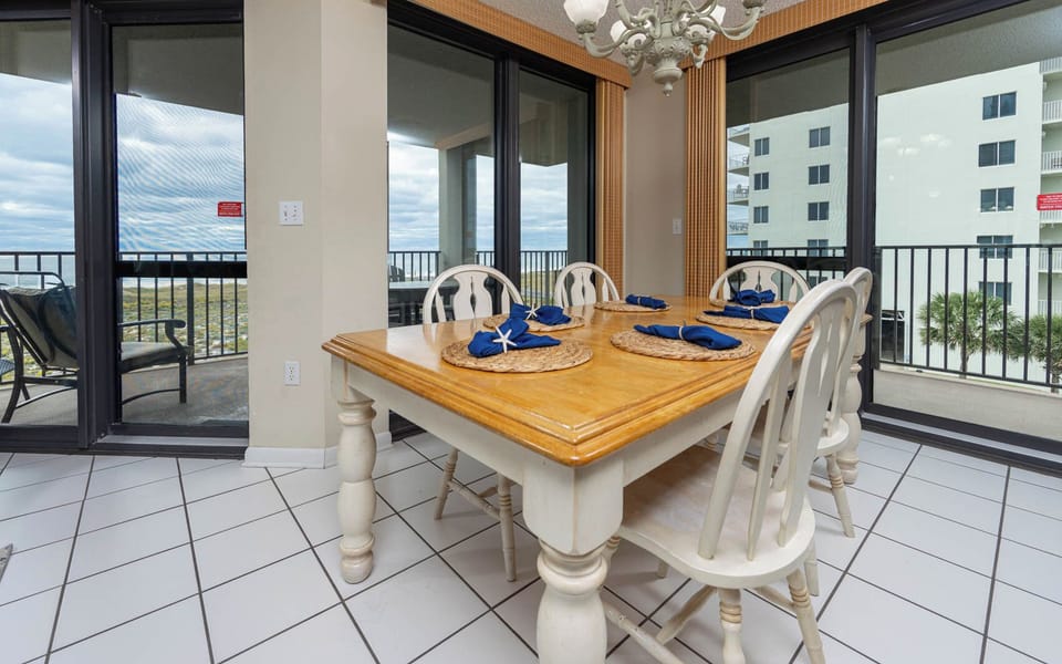 Dining area features a wooden table set for six with blue napkins, adjacent to sliding glass doors leading to a balcony