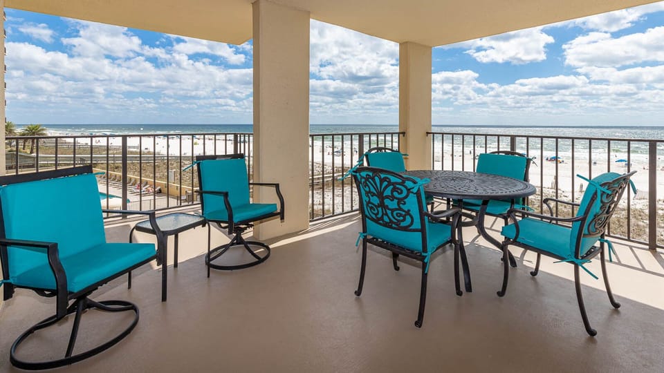 The balcony features turquoise chairs and a table, offering a view of the beach and gentle waves under a partly blue sky