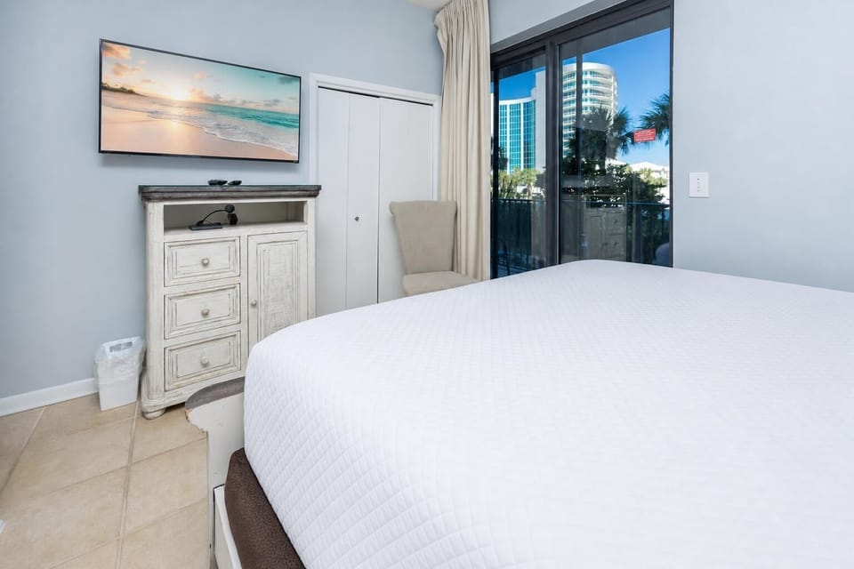 Relax in this comfortable guest bedroom featuring a large TV, a cozy chair, and natural light from the balcony doors