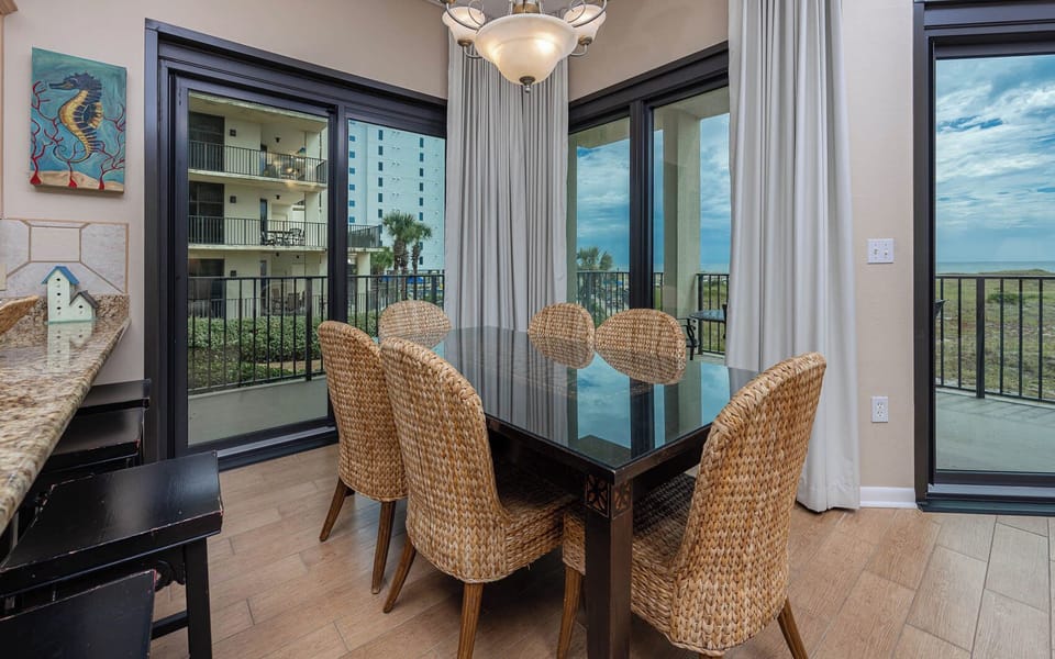 Dining area features a glass-top table surrounded by woven chairs, with large windows offering a glimpse of the beach