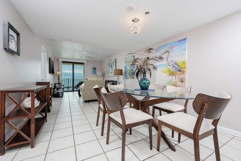 The living area features a glass dining table, cozy seating, and a breathtaking beach view through large sliding doors