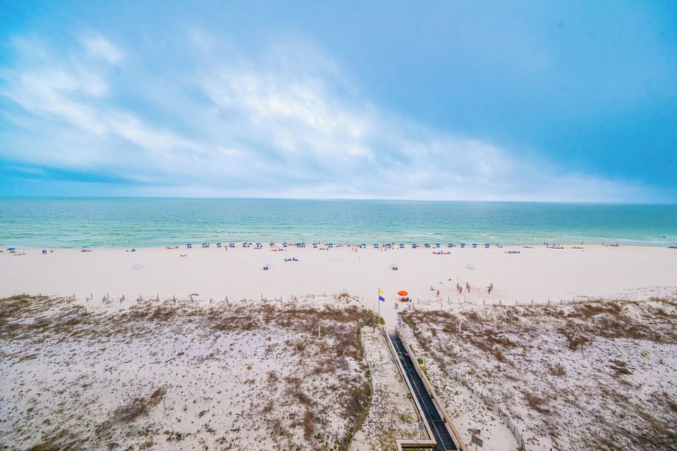 Dunes frame the walkway leading to the coast where families gather beneath colorful umbrellas on the white sand beach