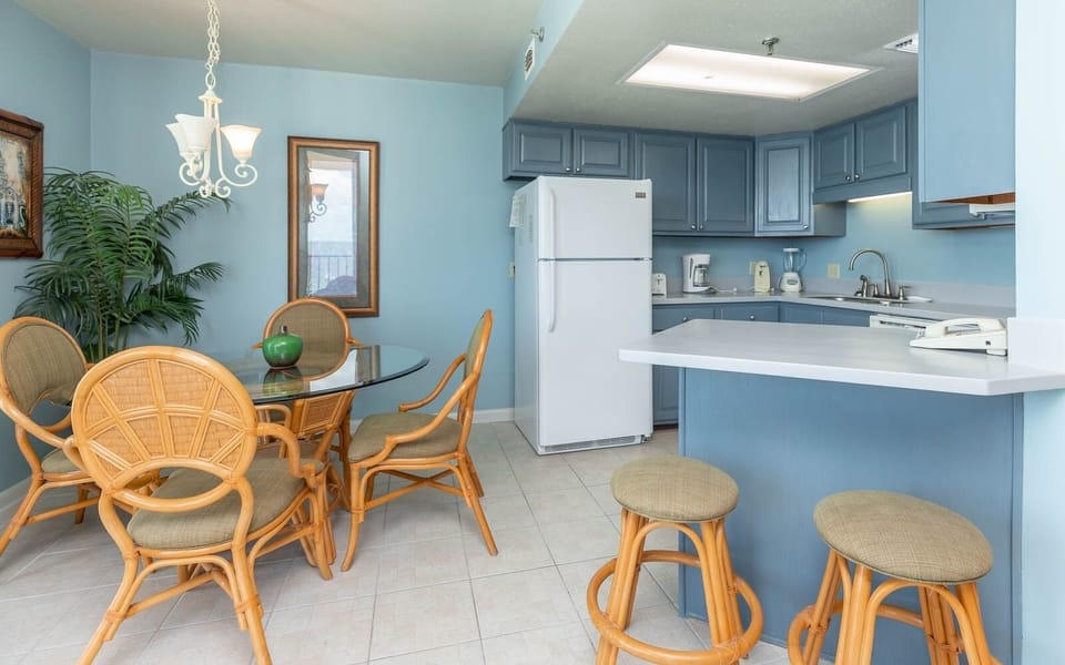 The bright kitchen features blue cabinetry, a round glass dining table with rattan chairs, and a convenient breakfast bar
