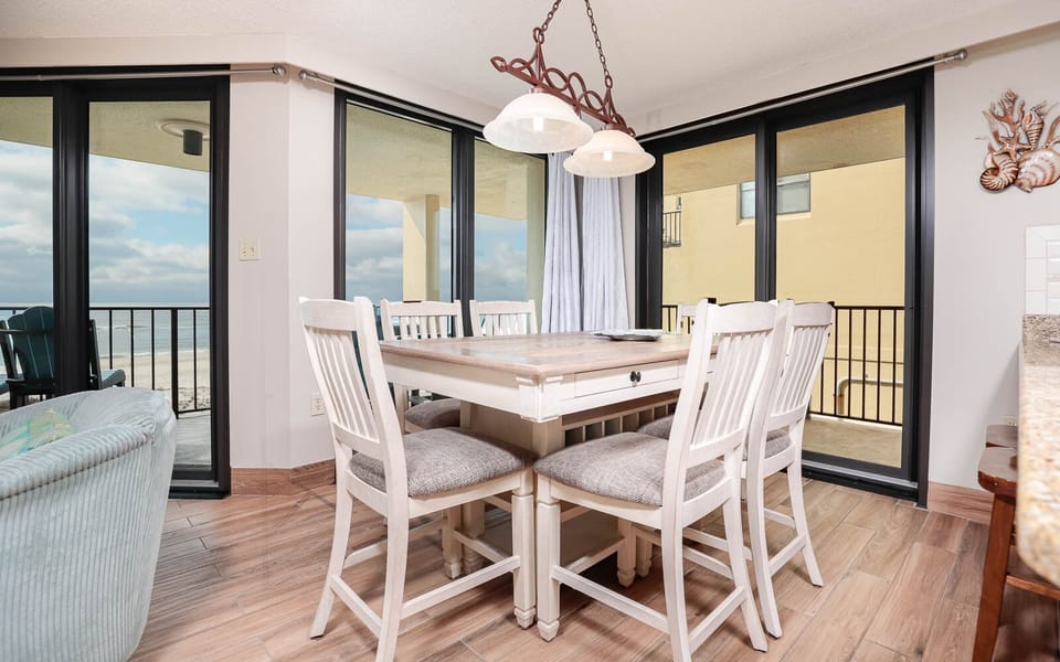 The dining area features a wooden table surrounded by cushioned chairs, with large windows showcasing a beach front view
