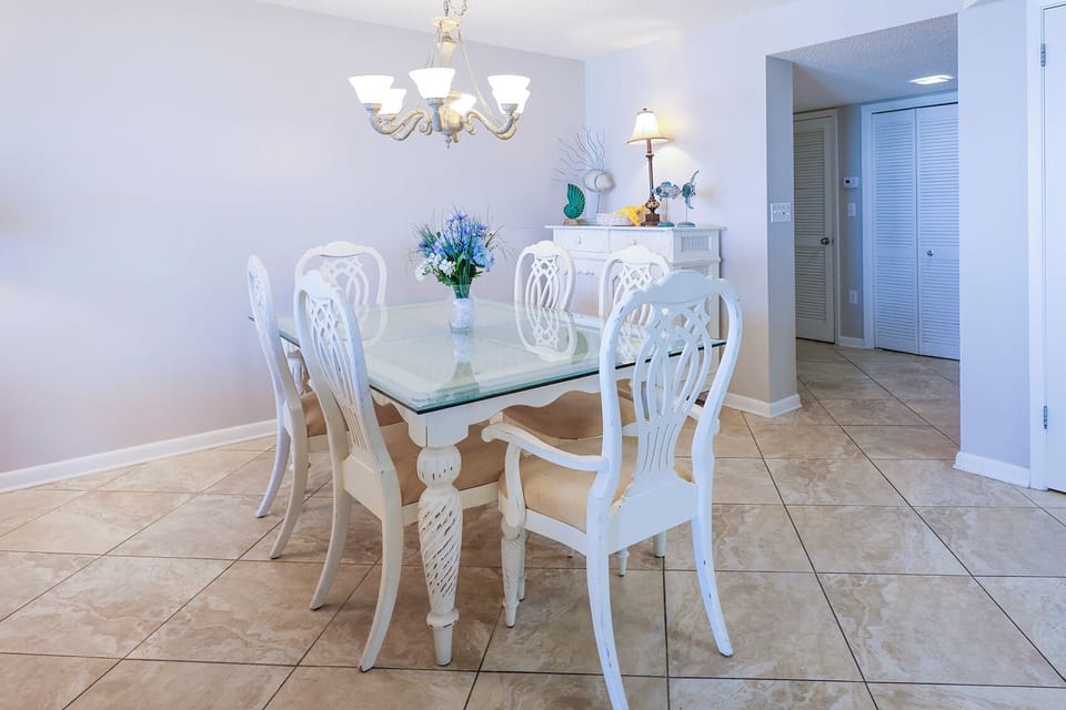 Bright dining area features a glass-top table surrounded by elegant white chairs, complemented by a charming chandelier