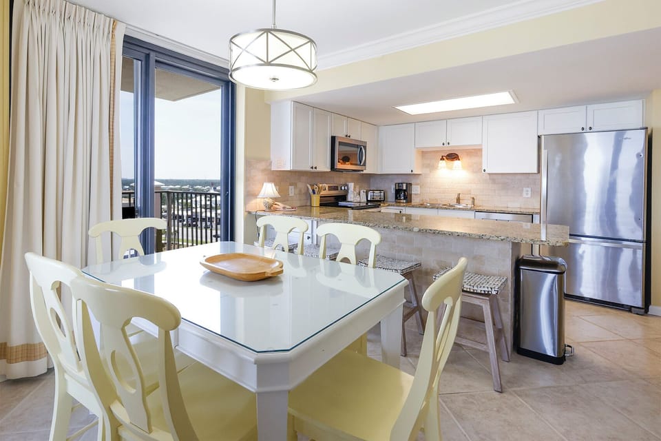 Dining space featuring a glass-top table with yellow chairs, complemented by a modern kitchen and access to the balcony