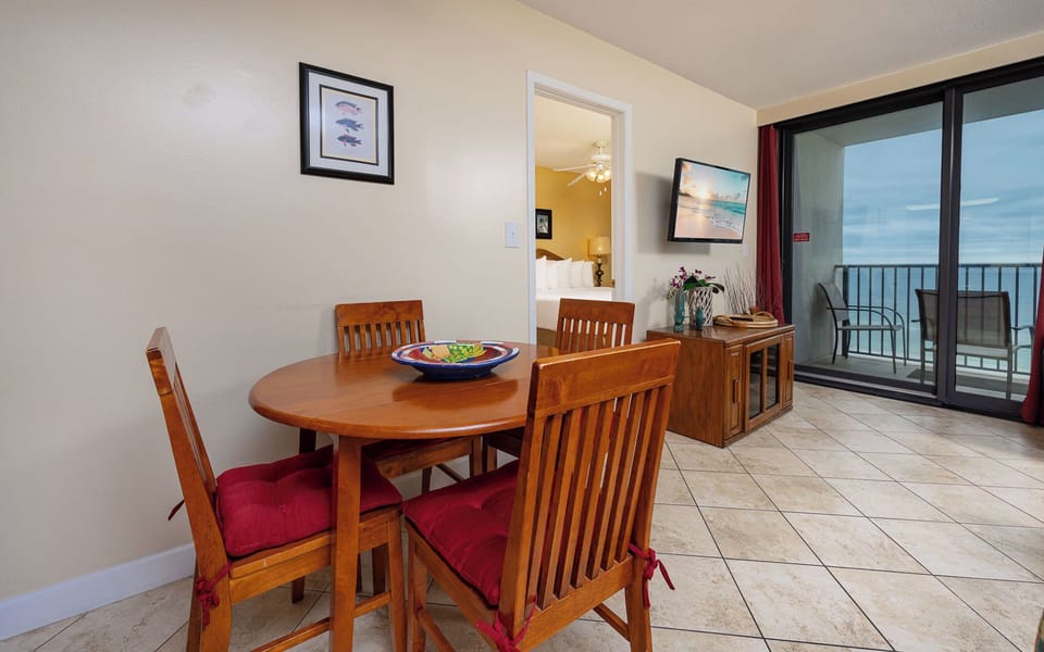 Bright dining area with a wooden table, red cushioned chairs, and a view of the balcony overlooking the serene coastline