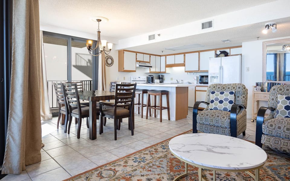 The open living area features a dining table, breakfast bar, and cozy seating with beach views visible through large windows