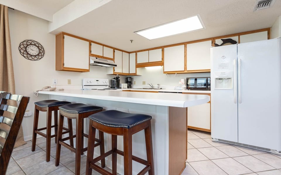 The kitchen features light wood cabinetry, a spacious countertop, Keurig, coffee maker, and bar stools with navy cushions