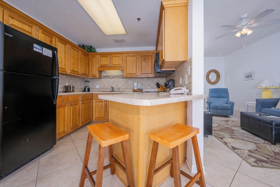 The kitchen features warm wood cabinetry, a breakfast bar with two stools, and a view into the cozy living area