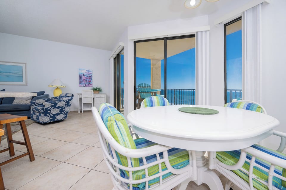 Bright dining area features a round table with colorful striped chairs, offering a view of the beach through large windows