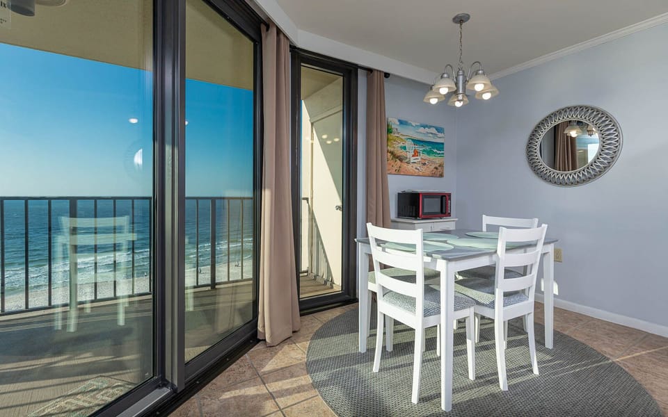 Bright dining area features a round rug, white table with four chairs, and a view of the beach through large glass doors