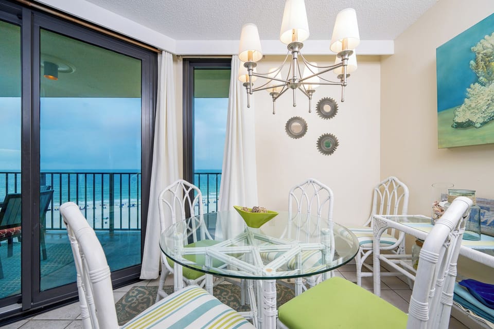 Bright dining area featuring a glass table and white chairs, with expansive views of the beach through large windows