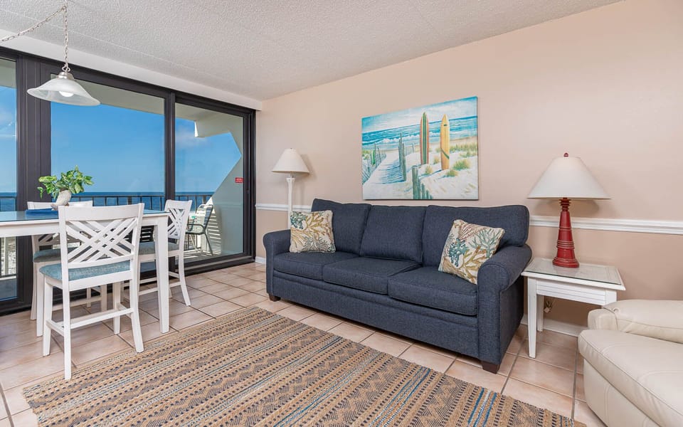 The living room features a comfortable couch, a dining table, and stunning views of the beach through large windows