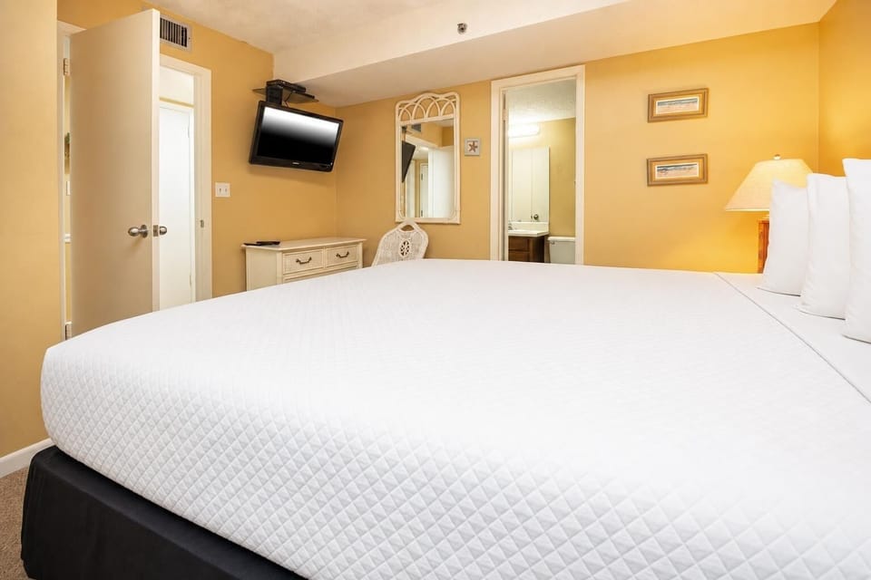 This cozy bedroom features a plush king bed, a mounted TV, and warm yellow walls, creating a relaxing retreat
