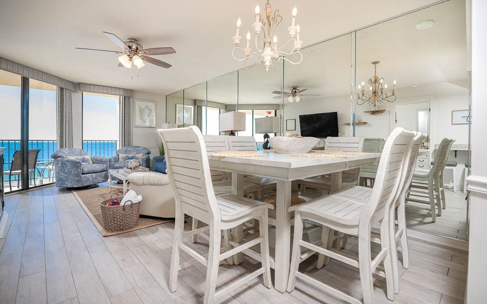 Bright dining area with white wooden furniture, a chandelier, and views of the beach through large windows