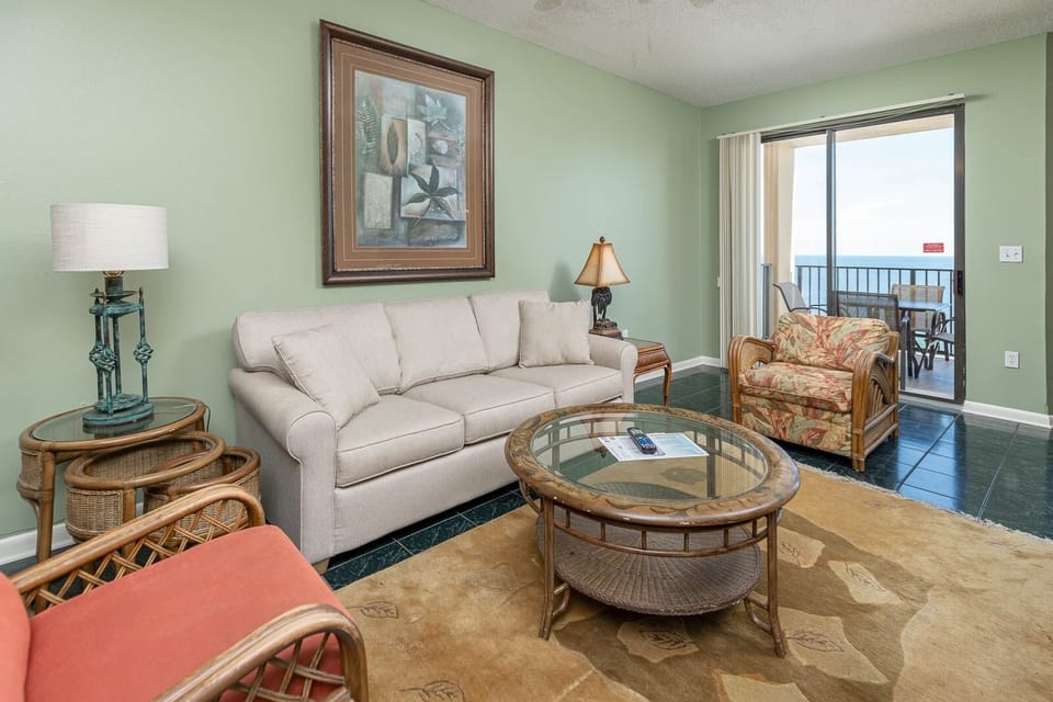 A light-filled living area features a beige sofa with queen sleeper, armchair, and balcony doors leading to a beach view