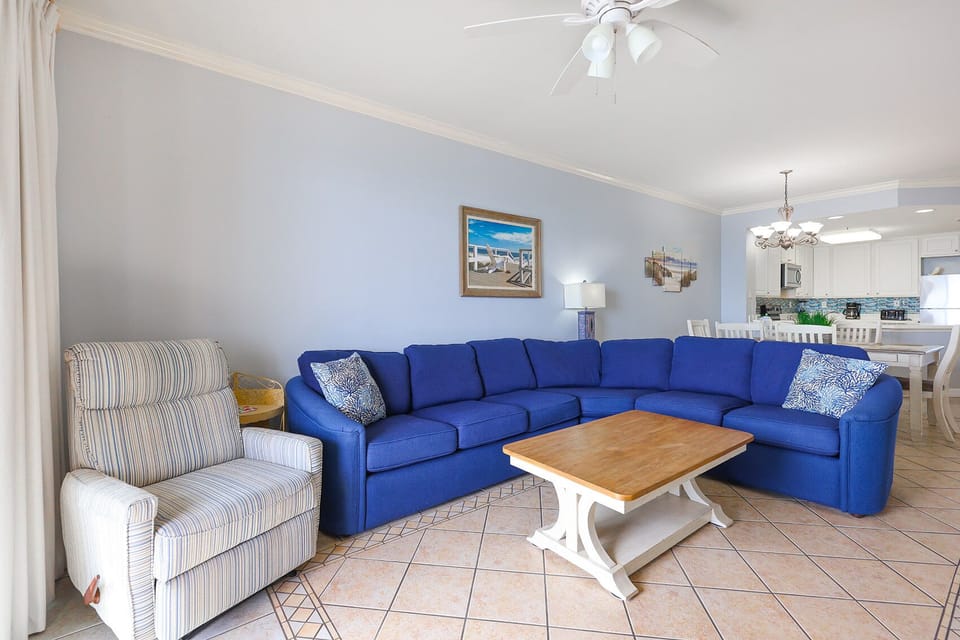 The living area features a vibrant blue sectional sofa, a wooden coffee table, and a view of the kitchen with bright cabinets