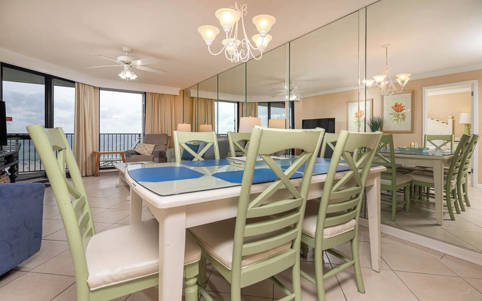 Dining area features a glass-top table with green chairs, overlooking the beach through large windows and soft curtains