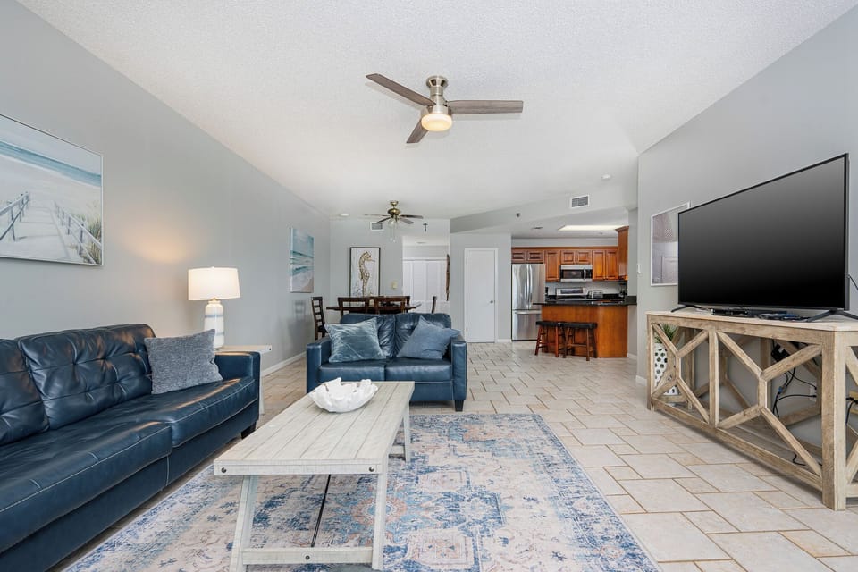 Living area featuring a dark blue sofa with queen sleeper, light wood coffee table, and a large TV, leading to the kitchen
