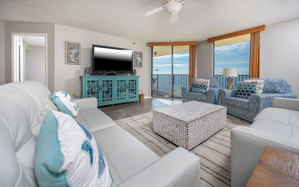 Bright living area features a white leather sofa, blue accent chairs, and a large TV, with a view of the beach beyond