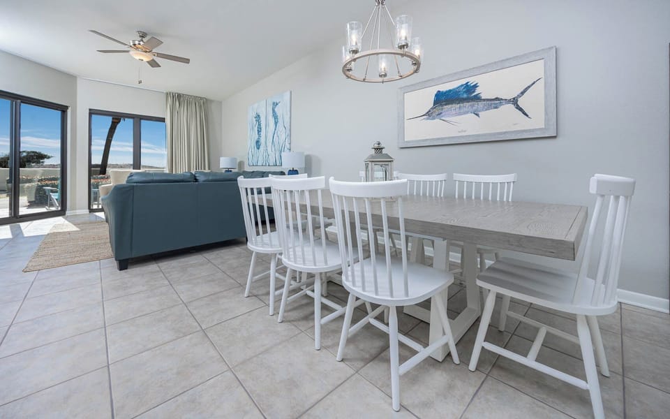 Spacious dining area features a large wooden table with white chairs, complemented by coastal artwork and natural light