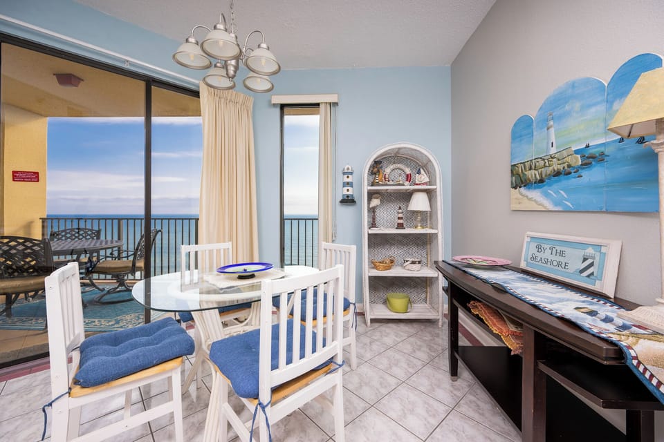 Bright dining area features a glass table with blue-cushioned chairs and a coastal decor, offering a view of the beach