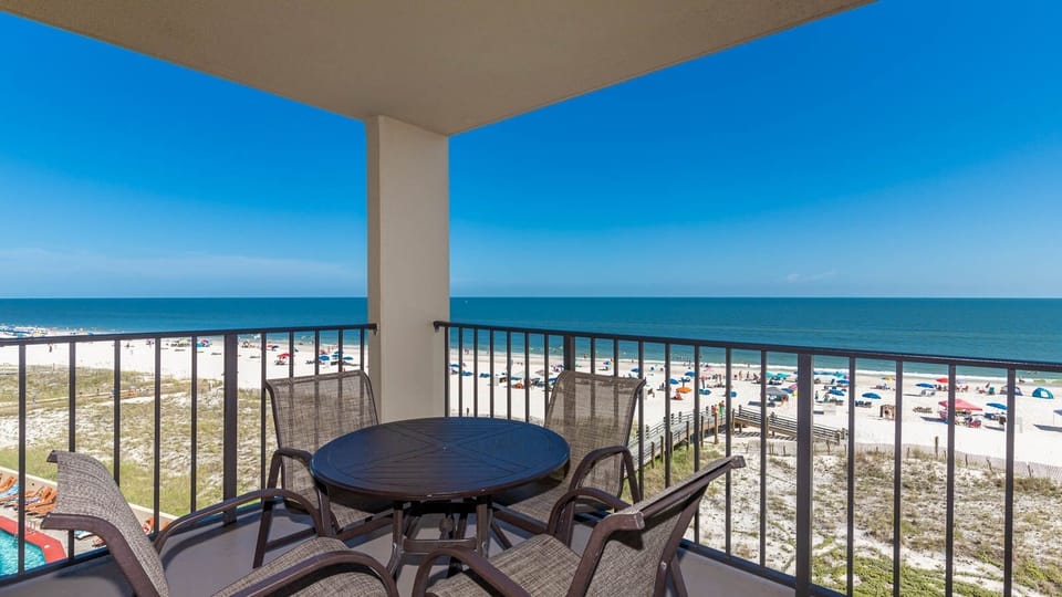 The balcony features a round table and chairs, offering a clear view of the beach and vibrant umbrellas dotting the shore