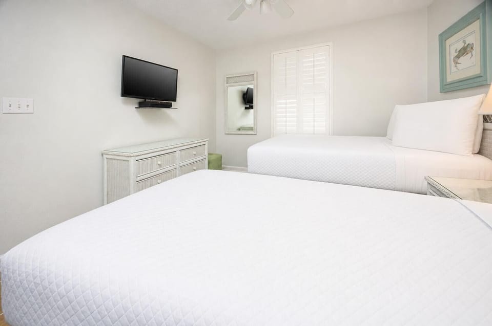 Bright guest bedroom featuring two double beds, a wall-mounted TV, and a dresser, complemented by natural light