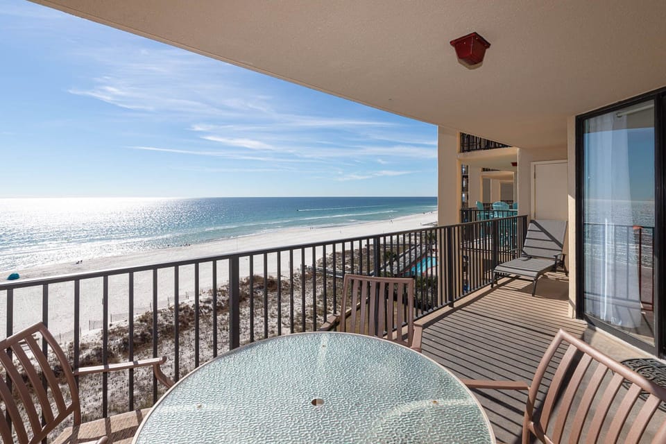 The balcony features a glass-top table and chairs, offering a serene view of the beach and sparkling water beyond