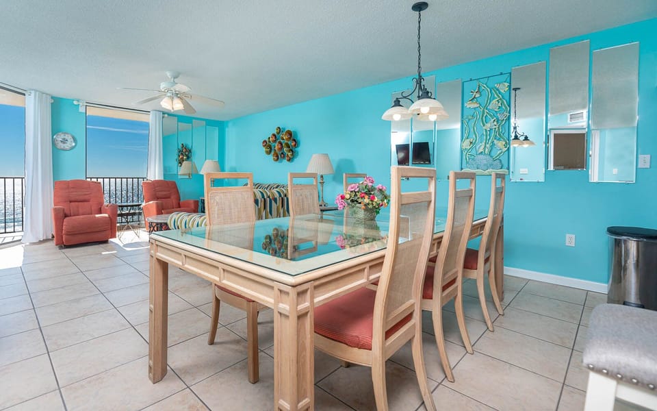Bright turquoise walls complement a dining area with a glass-top table, surrounded by six chairs and views of the beach