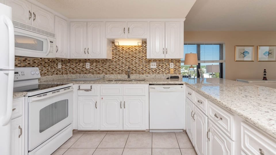 This kitchen features sleek white cabinetry, granite countertops, and a view of the beach, perfect for culinary adventures
