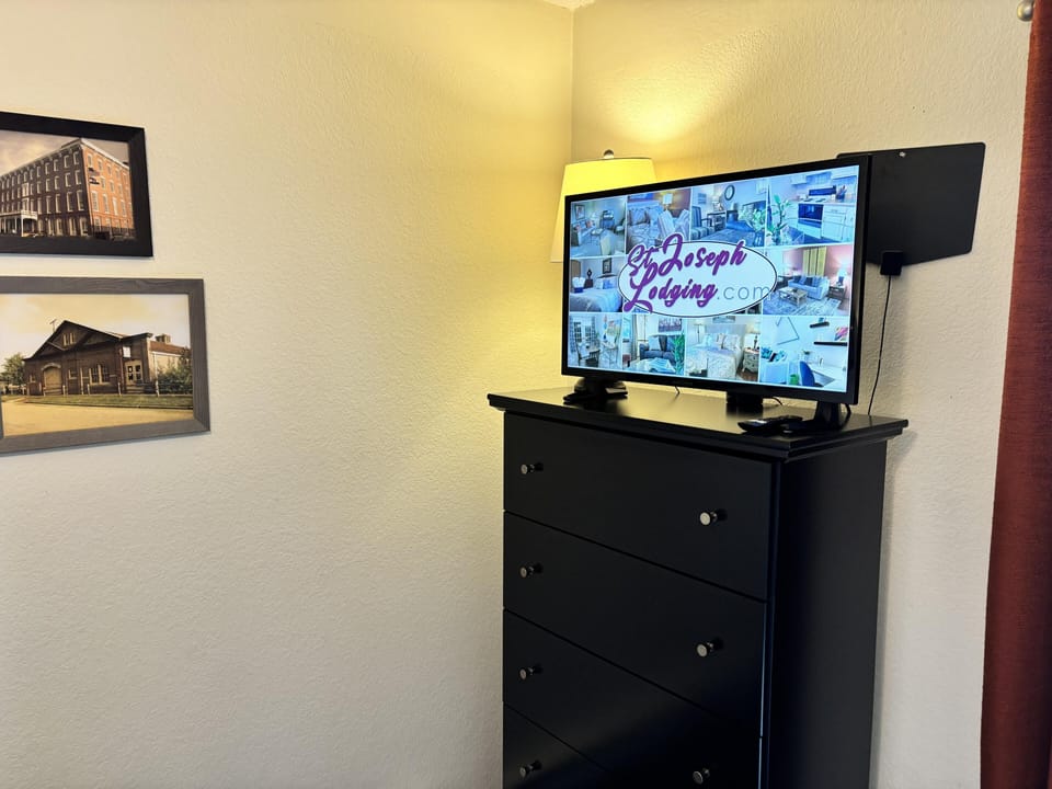 Flat-screen TV sits atop a tall black dresser, illuminated by a nearby floor lamp in the bedroom corner.
